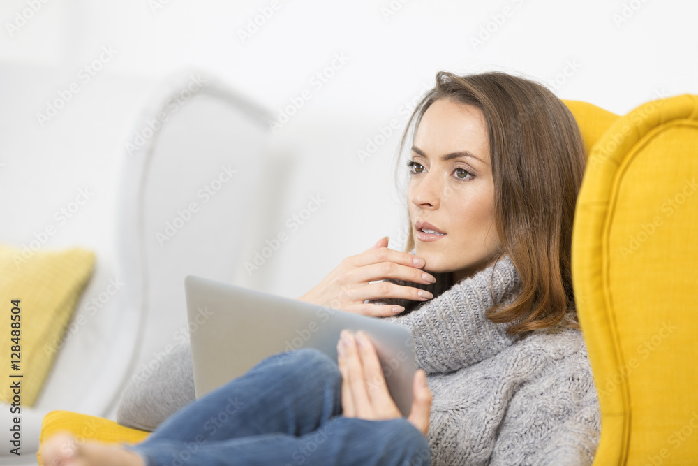 Woman using her laptop at home in a yellow chair relaxing in winter clothes