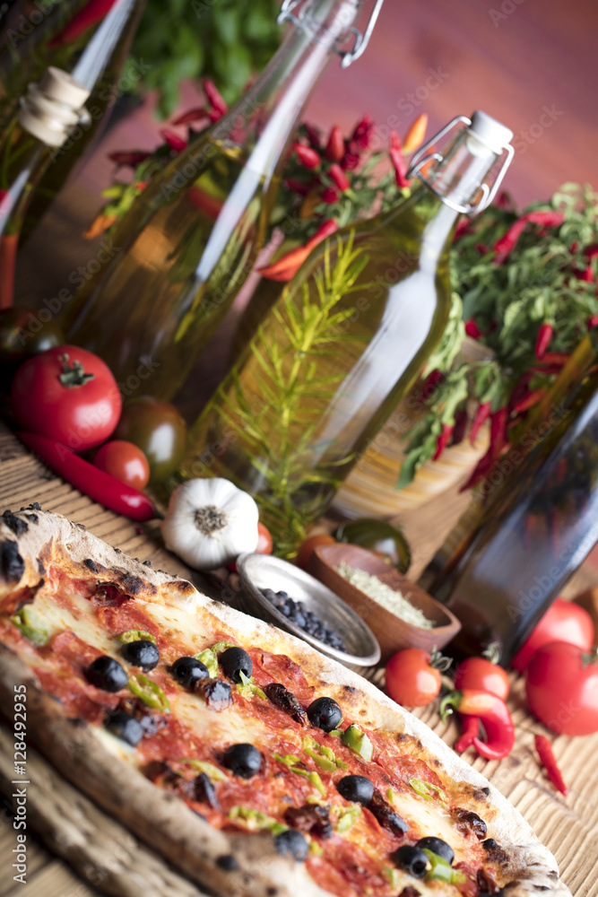 Delicious, tasty and fresh, rustic Italian pizza, served on wooden table. Bunch of tomatoes, chili, basil, garlic in background. Bowls with different kinds of pepper. Many bottles of olive oil