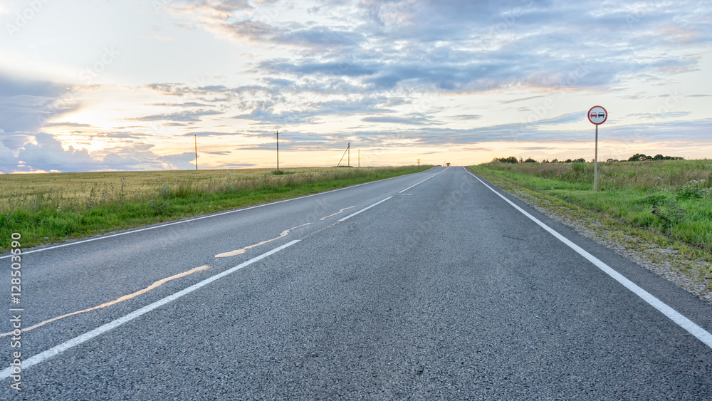 Vanishing straight highway through meadow at sunset in back lit. Silkovo, Kaluzhsky region, Russia.
