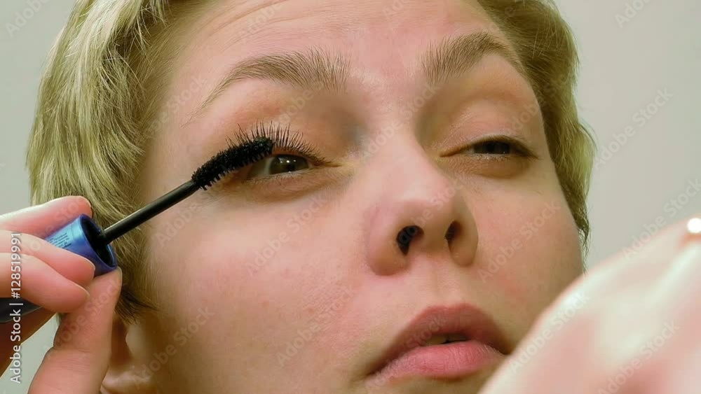 Pretty blond woman applying mascara make-up on her eyelashes in front of mirror and looks at herself. Beauty and makeup concept.