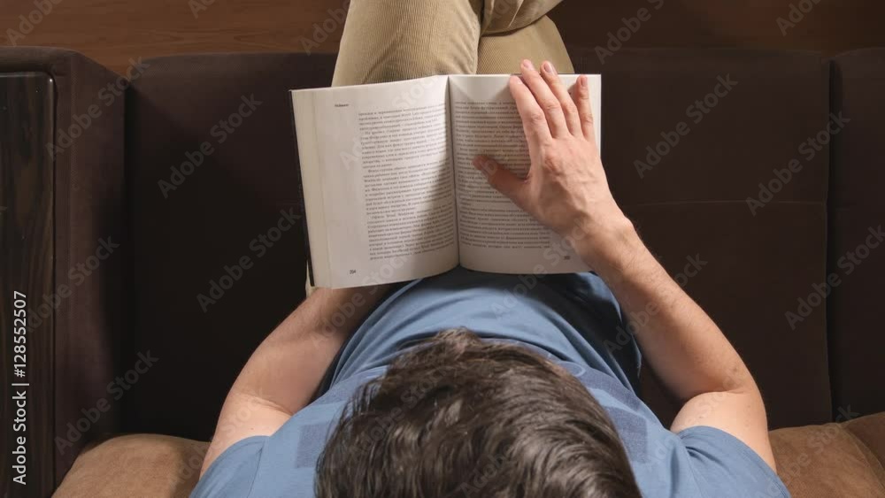 TOP VIEW: Young adult man reads a book on a sofa at home TOP VIEW: Young adult man reads a book on a sofa at home