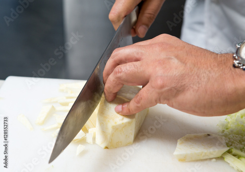 A knife cutting thin cubes of fresh organic celeriac root vegetable, on a white cutting board.