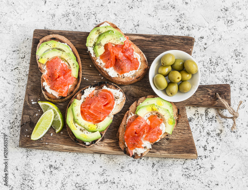 Delicious appetizers - cream cheese, smoked salmon and avocado sandwiches and olives on a wooden board. On a light background, top view