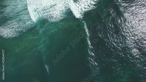 Forward flight over the ocean looking straight down at waves crushing with force on rocks.