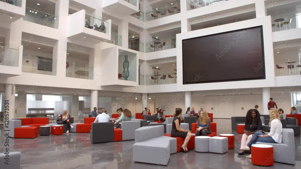 Students sit talking under AV screen in atrium at university