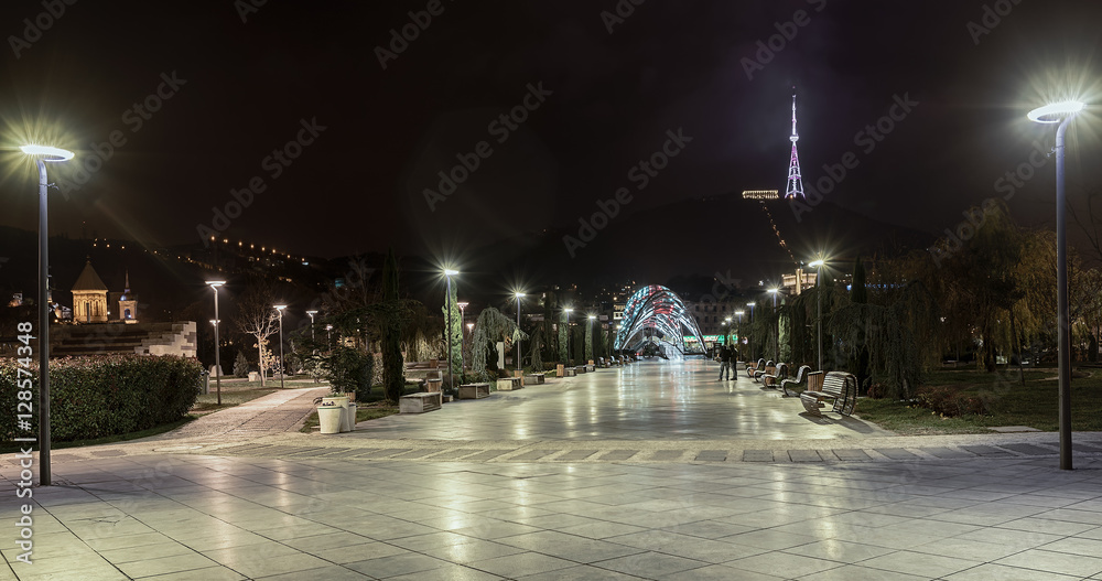 Fototapeta premium Georgia, Tbilisi - November 23, 2016. Panorama of the night city. Recreation Park Rijk, TV tower, pedestrian bridge Peace empty benches and light on the lampposts.
