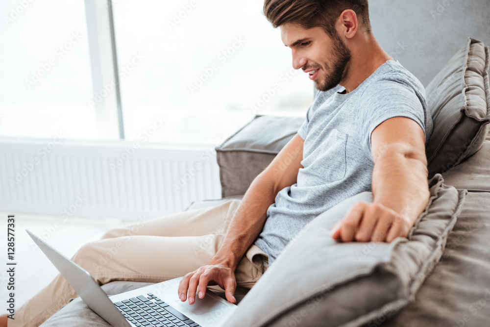 Cheerful young man sitting on sofa and working by laptop