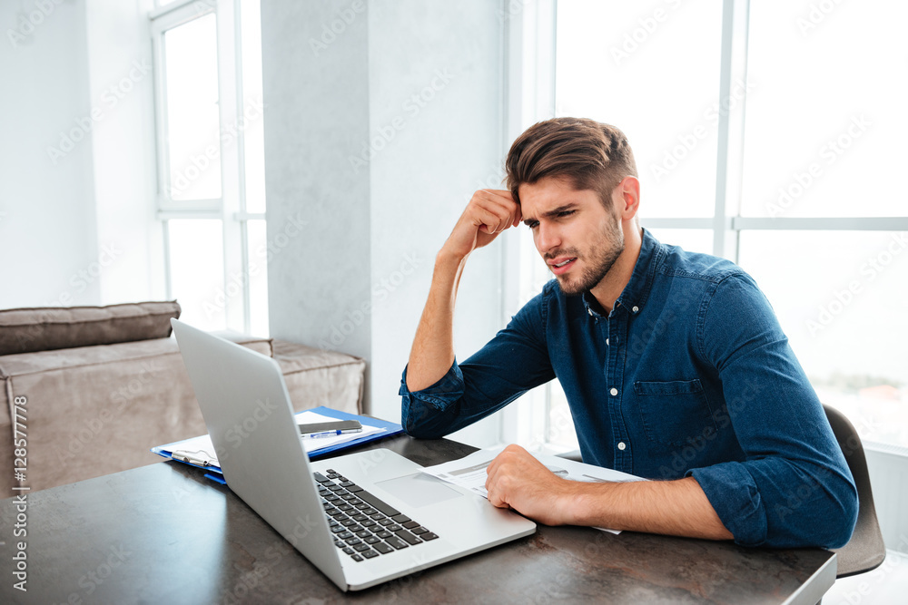 Sad young man sitting near laptop and holding head Sad young man sitting near laptop and holding head