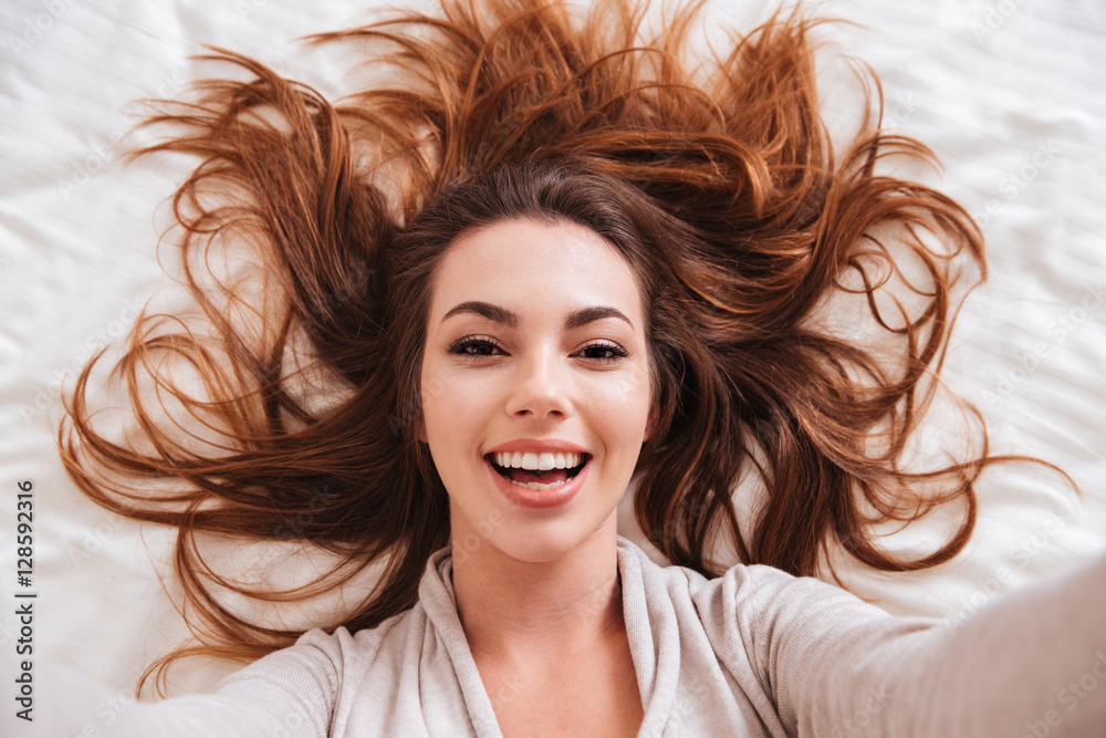 Woman with beautiful hair lying in bed and taking selfie