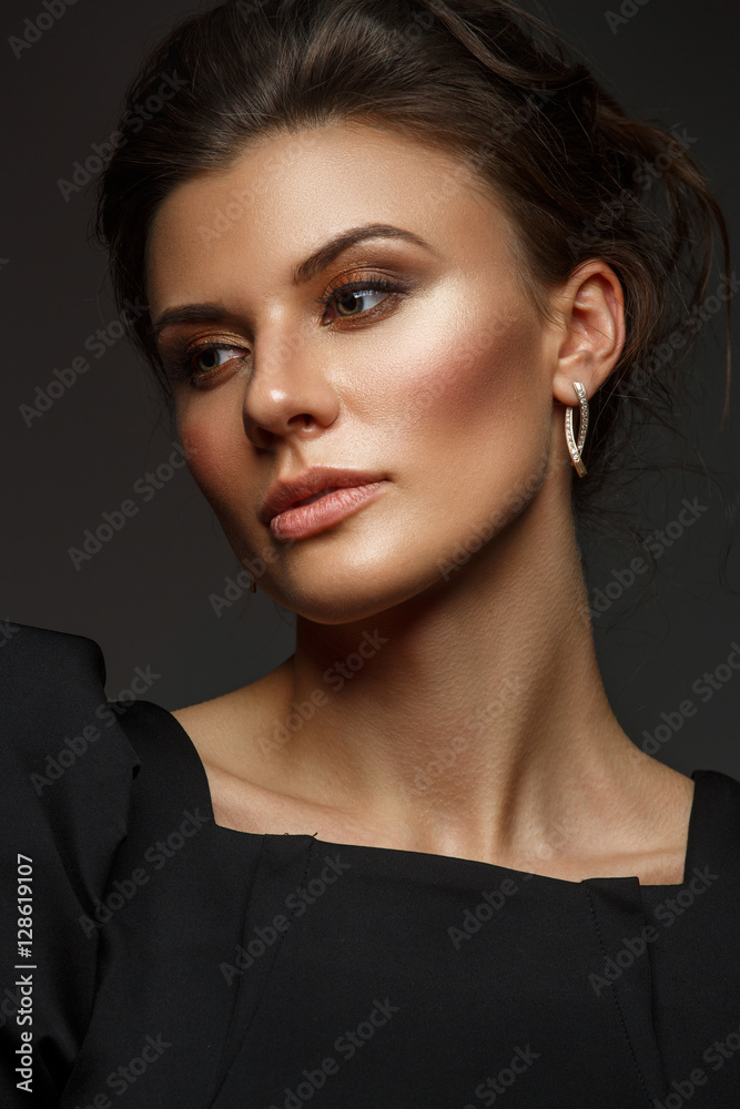 Fototapeta premium Portrait of a beautiful, self-sufficient and confident young woman with collected hair in the elegant black dress with makeup, dark background, studio photo
