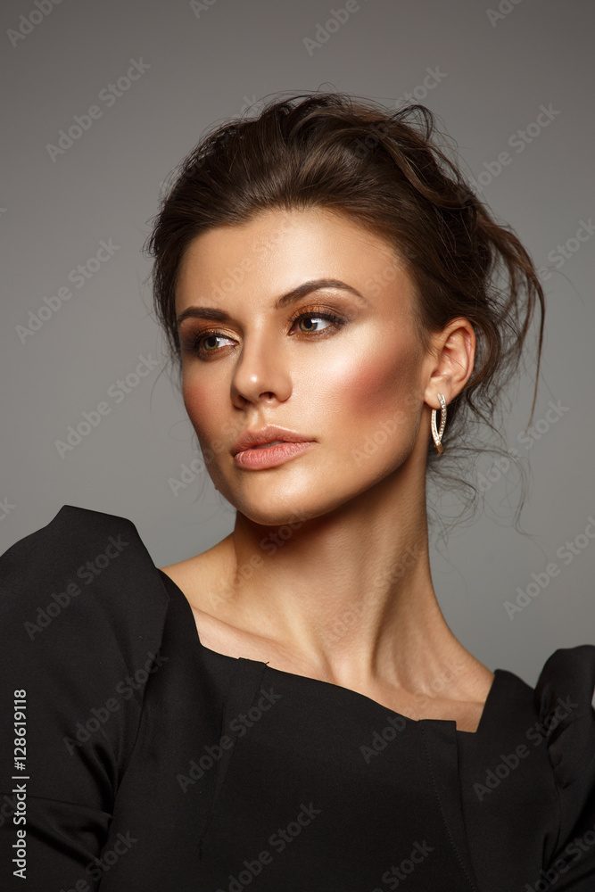 Fototapeta premium Portrait of a beautiful, self-sufficient and confident young woman with collected hair in the elegant black dress with makeup, dark background, studio photo