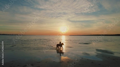 Two GIrls are Riding Horses on a Beach. Horses Walk on Water. Beautiful Sunset is Seen in this Aerial Shot.