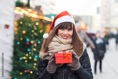 beautiful woman in santa hat holding present box on christmas market