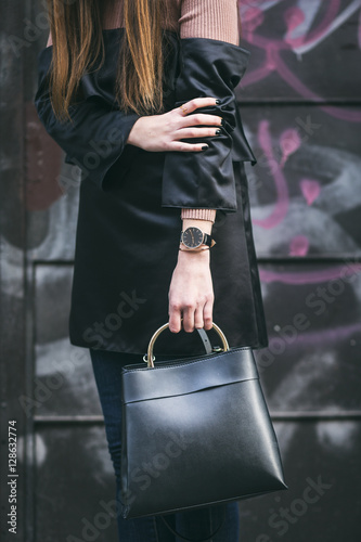 stylish woman wearing a black, silk off the shoulder dress and a light pink sweater. accessorized with a black and golden watch and an elegant black handbag with a golden handle.