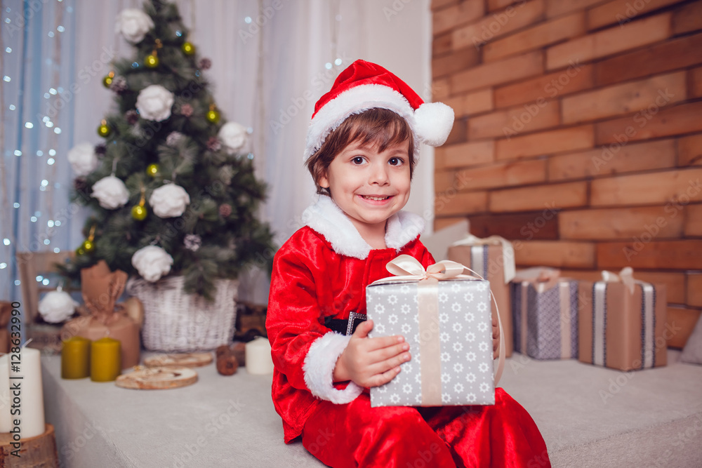 Little boy dressed as Santa Claus sitting with gift in Christmas decoration. New Year. Little boy dressed as Santa Claus sitting with gift in Christmas decoration. New Year.
