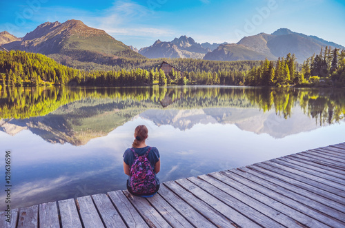 Alone young girl sitting and resting on the wooden path near by beautiful blue lake and clear big mountains. Original wallpaper from summer morning