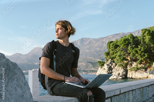 Charming Young man working on laptop near the sea, freelance wor