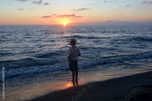 Kleiner Junge steht im Wasser am Strand beim Sonnenuntergang und schaut zur Sonne
