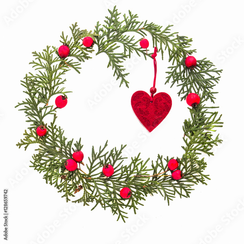 Christmas frame made of green thuja twigs, red wild rose fruits and Xmas tree decoration felt heart on white background. Top view, flat lay. Copy space for text