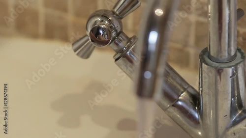 A shot of man turning the tap and washing his hands beneath the flowing water under a shining silver tap and tiled background.