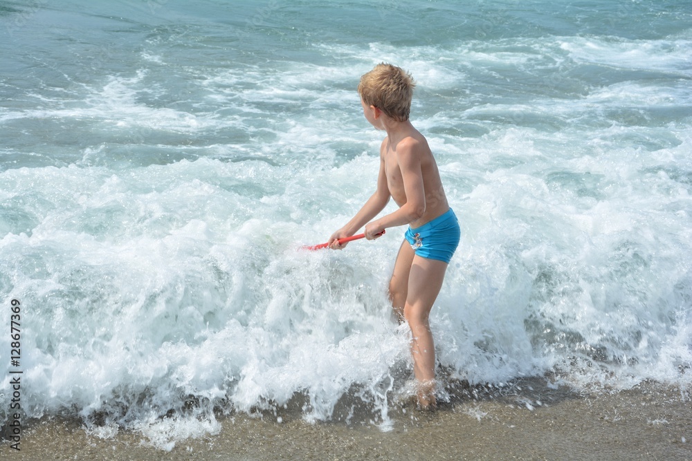 Kleiner blonder Junge mit blauer Badehose steht mit einem Kescher direkt in der Meeres - Welle Kleiner blonder Junge mit blauer Badehose steht mit einem Kescher direkt in der Meeres - Welle