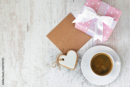Gift box wrapped in pink dotted paper, heart shaped love cookie, a cup of coffee and an empty kraft card over a white wood background.
