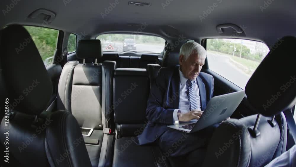 Fish eye shot of senior businessman sitting in moving car talking on phone and working on his laptop Fish eye shot of senior businessman sitting in moving car talking on phone and working on his laptop