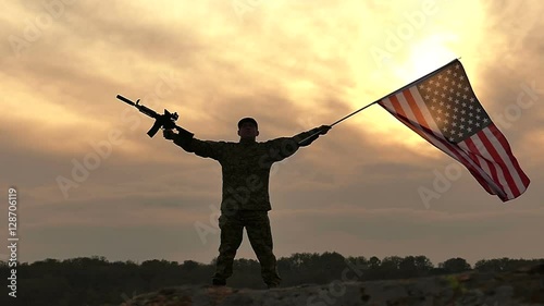 
Soldier stand with American flag and rifle against gloomy orange  sky. Slow Motion
