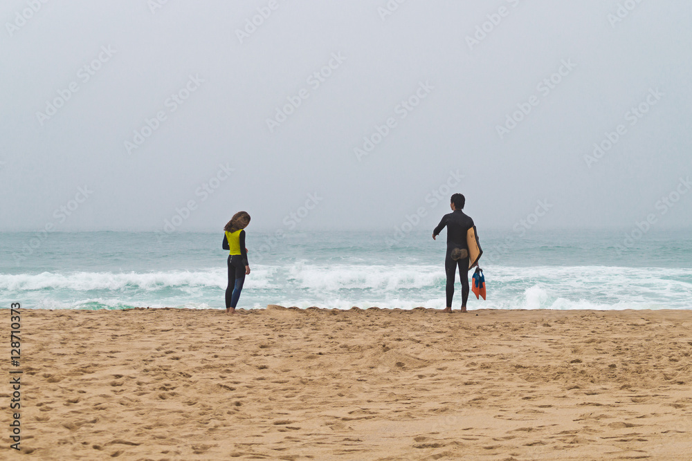 Two surfers stand ashore ocean. Fellow and girl.