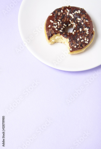An iced chocolate sprinkle ring donut with bite mark, on a pastel purple background with blank space below