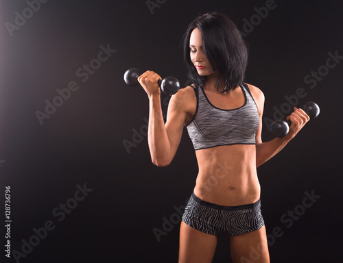 Bodybuilding concept. Sports woman training with dumbbells while demonstrating her muscular body isolated on black in studio.