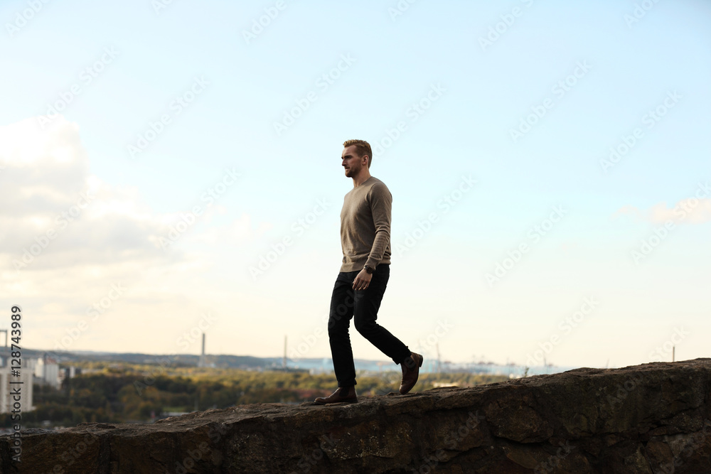 Man wearing a brown sweater and jeans, walking along a mountain top with the great cityview behind him on a summer day. Looking away from camera.