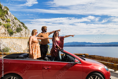 Group of happy young people waving and pointing fingers from the red convertible.