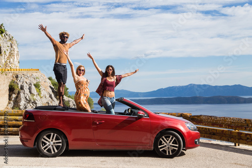 Group of happy young people standing in the red convertible car and waving.