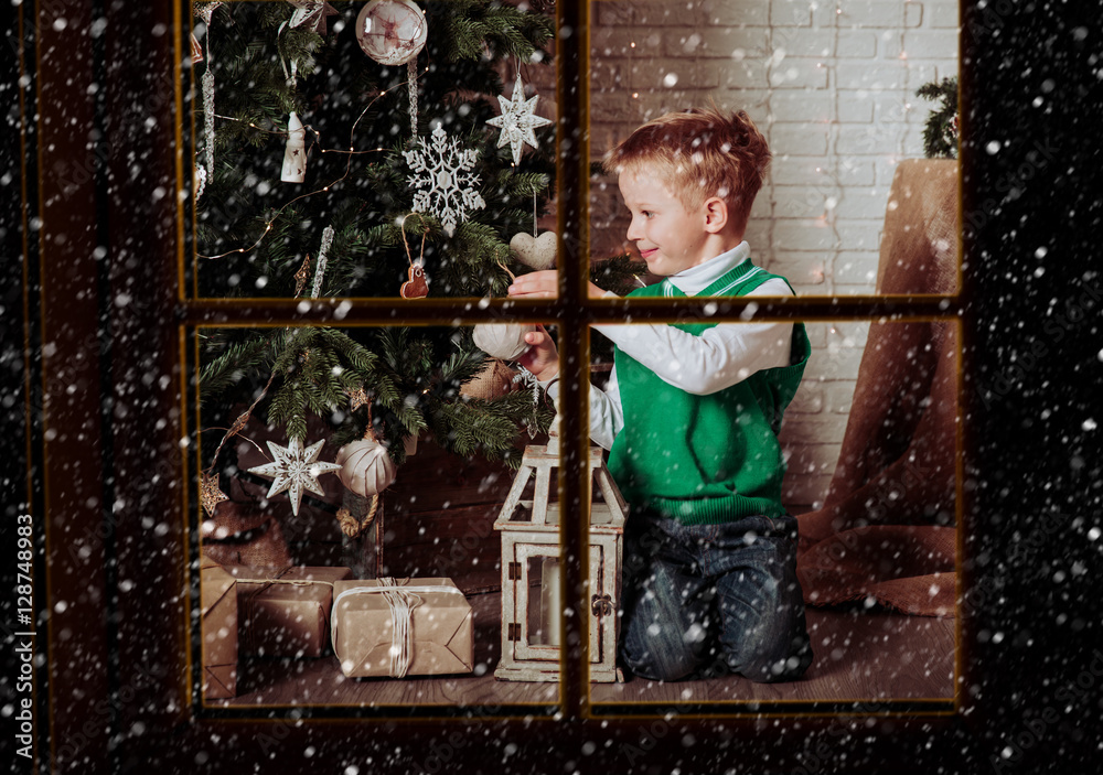little boy decorating Christmas tree viewed through window from outside