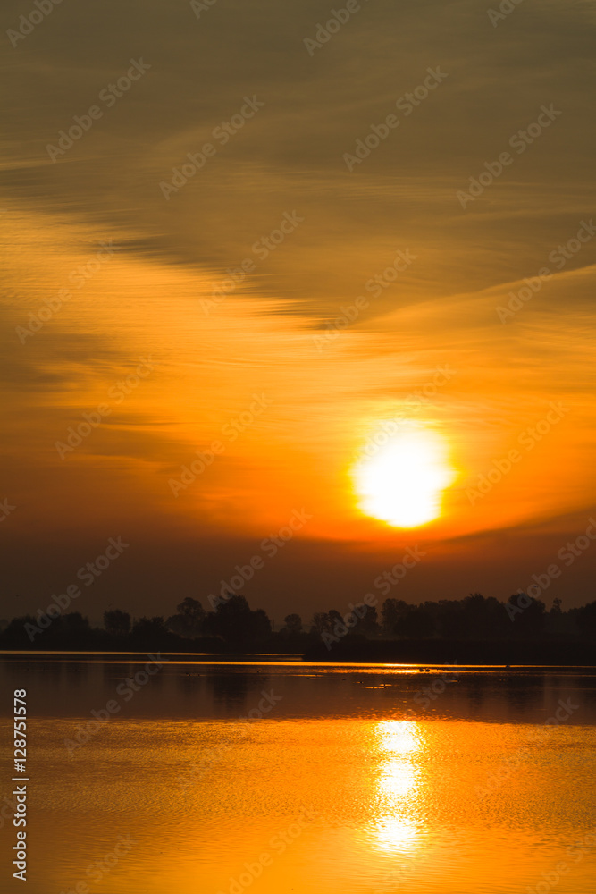 Sunrise over the surface water of lake with floating birds and curtain clouds in the red sky.