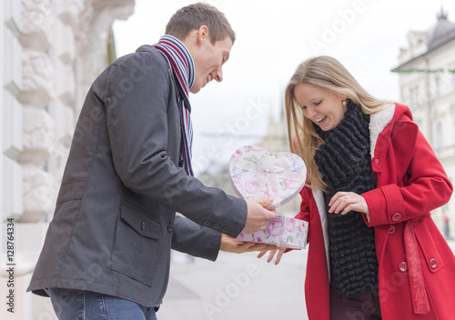 Fototapeta Naklejka Na Ścianę i Meble -  Handsome Young Man Giving His Girlfriend a Gift Box While Standing on the Street. Love and Valentines Day Concept.