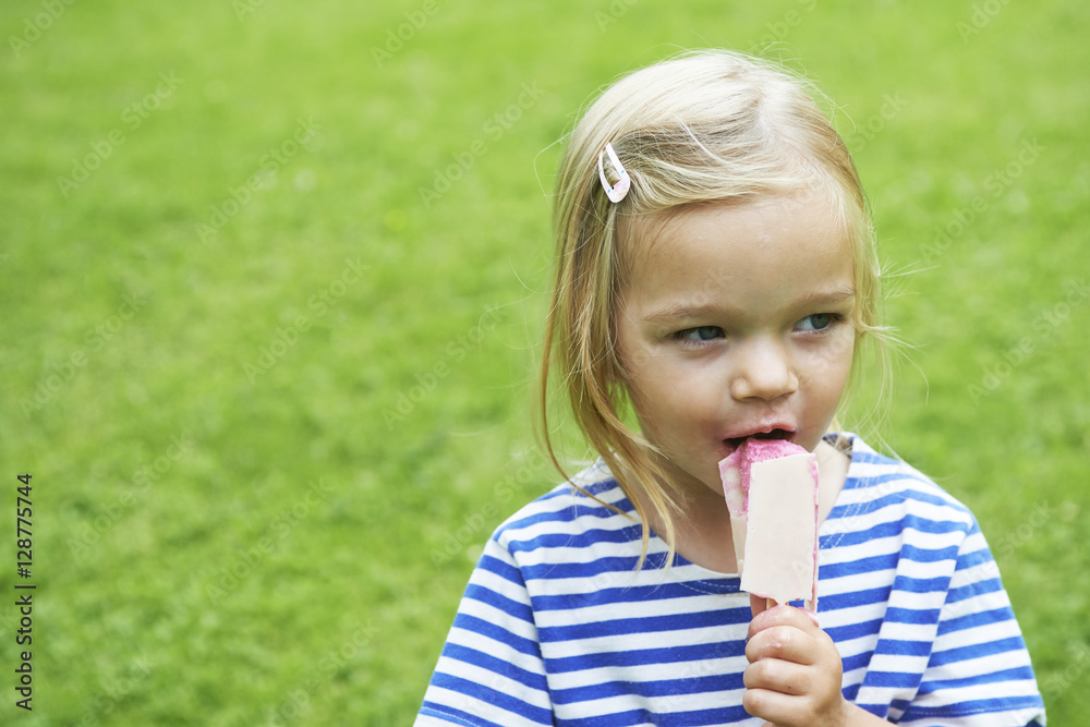 Beautiful little girl eats ice-cream in the summer with green grass background.
Beautiful little girl eats ice-cream in the summer with green grass background.