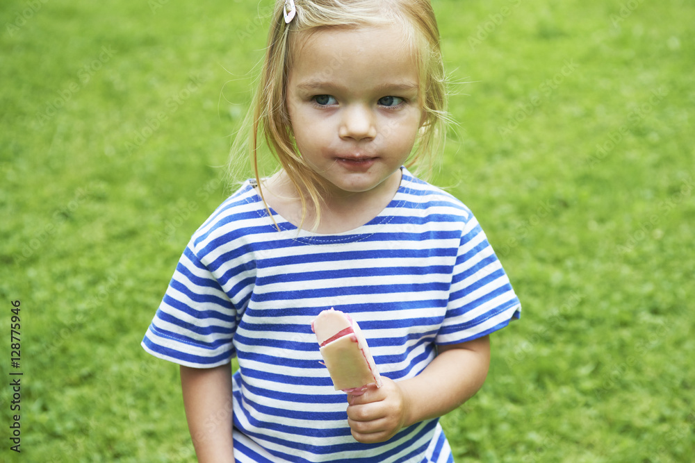 Beautiful little girl eats ice-cream in the summer with green grass background.
Beautiful little girl eats ice-cream in the summer with green grass background.