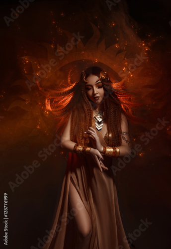 Beautiful, red-haired girl - a priestess of the sun. Dress and jewelry in ethnic style. Unusual make-up in orange and gold tones. Girl praying in silence. Fantastic photography