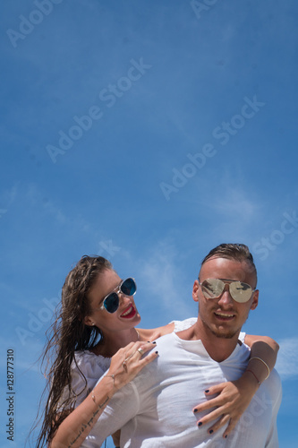 Cheerful handsome man carrying his beautiful girlfriend on his back on the beach over blue sky background