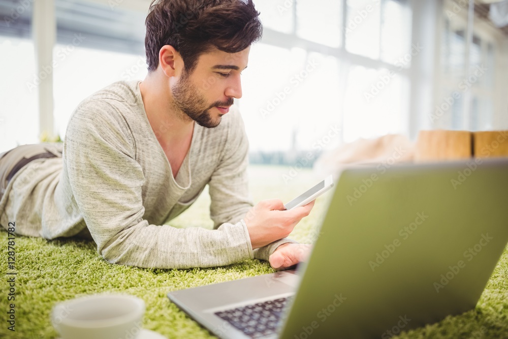 Businessman lying on carpet while using laptop and mobile phone Businessman lying on carpet while using laptop and mobile phone