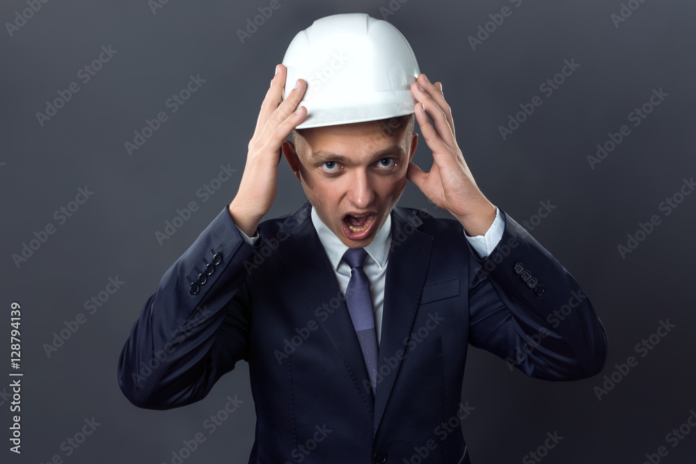 Handsome young engineer in a helmet he is holding his head and screaming standing on gray background Handsome young engineer in a helmet he is holding his head and screaming standing on gray background