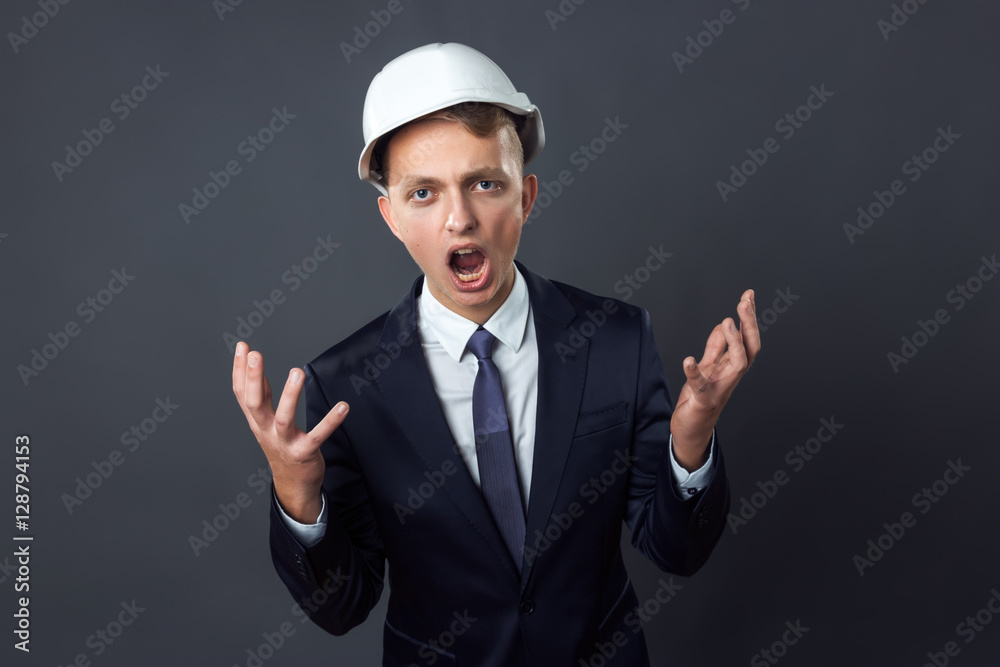 Handsome young engineer in a helmet he is screaming and spreads his arms standing on gray background