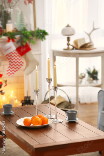 Wooden table with candles, cups and oranges in living room decorated for Christmas