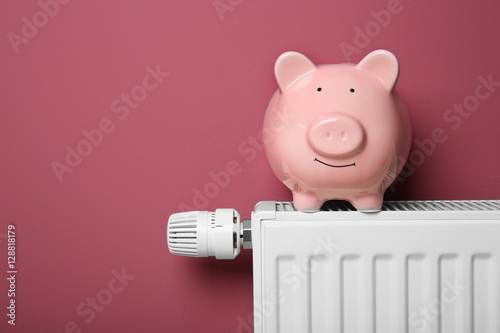 Piggy bank on heating radiator with temperature regulator on pink background, closeup