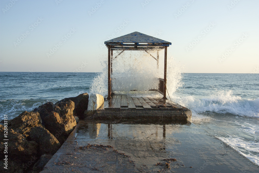 Gazebo on the rocky seashore is filled with a wave.