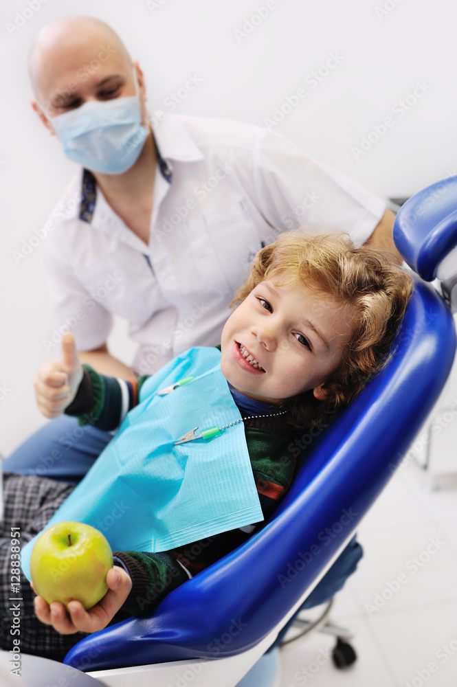 boy with curly red hair sitting in a dental chair and blue holding a green apple in hand. patient at the dentist's background