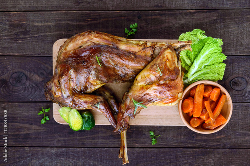 Roast whole rabbit on a wooden board with baked carrots and Brussels sprouts on a dark background. A festive meal. The top view.