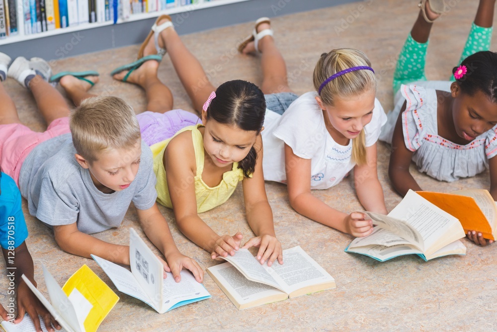 Fototapeta premium School kids lying on floor reading book in library 